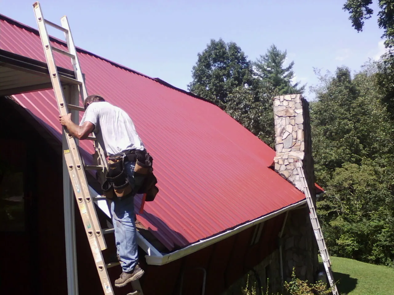 Roofer on a ladder beside a red metal roof and stone chimney — Soffit & Fascia Repair by Roofing Brothers in Oliver Springs